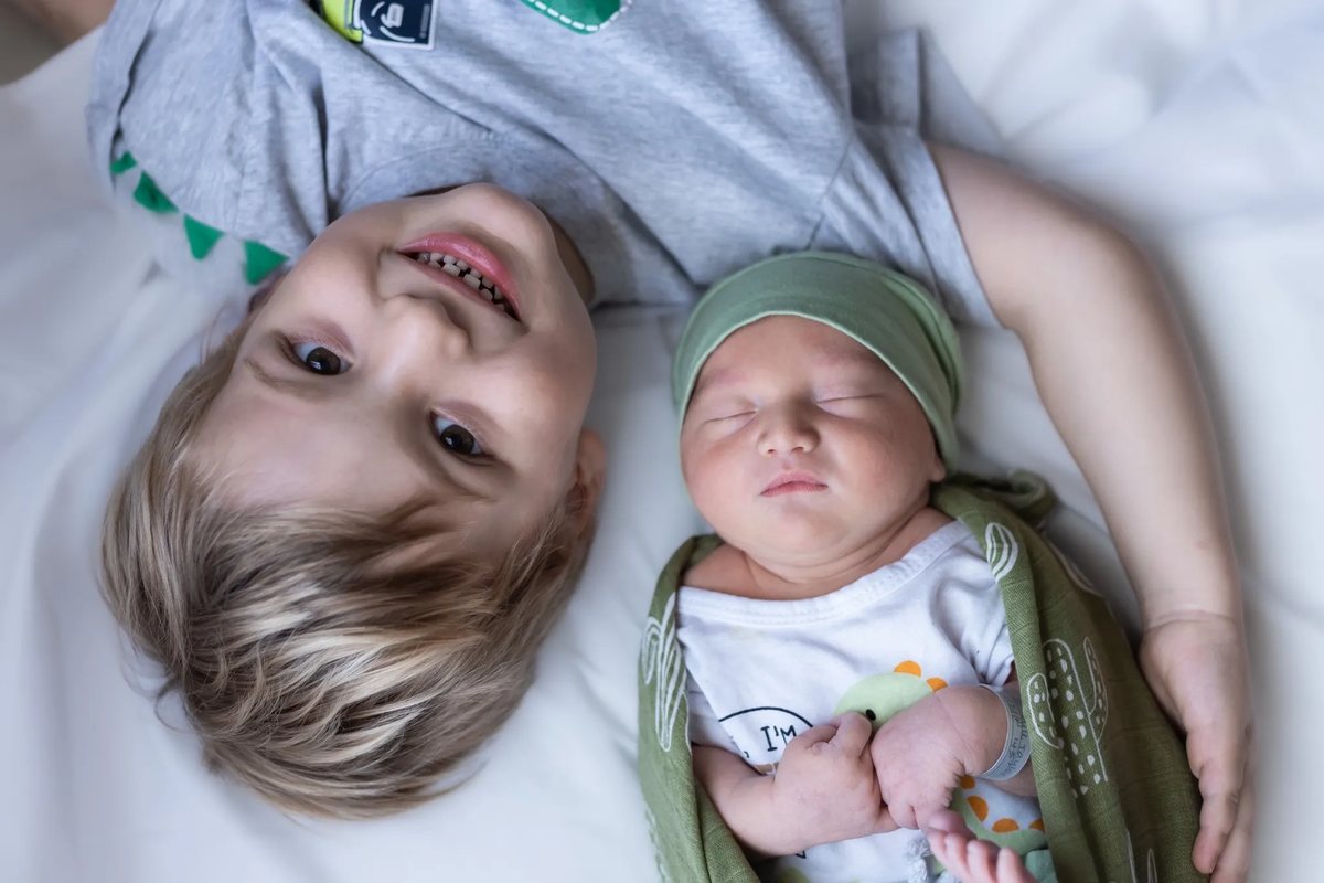A toddler and his newborn brother lying together on a white blanket