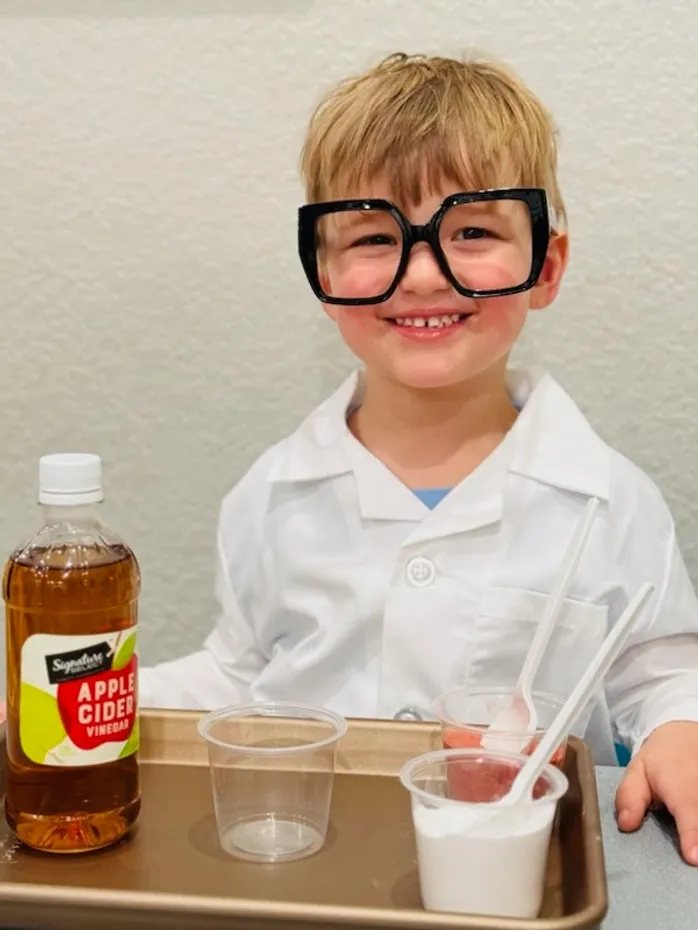 A young boy in oversized black glasses and a white lab coat grinning behind a tray of cups and a bottle of apple cider vinegar